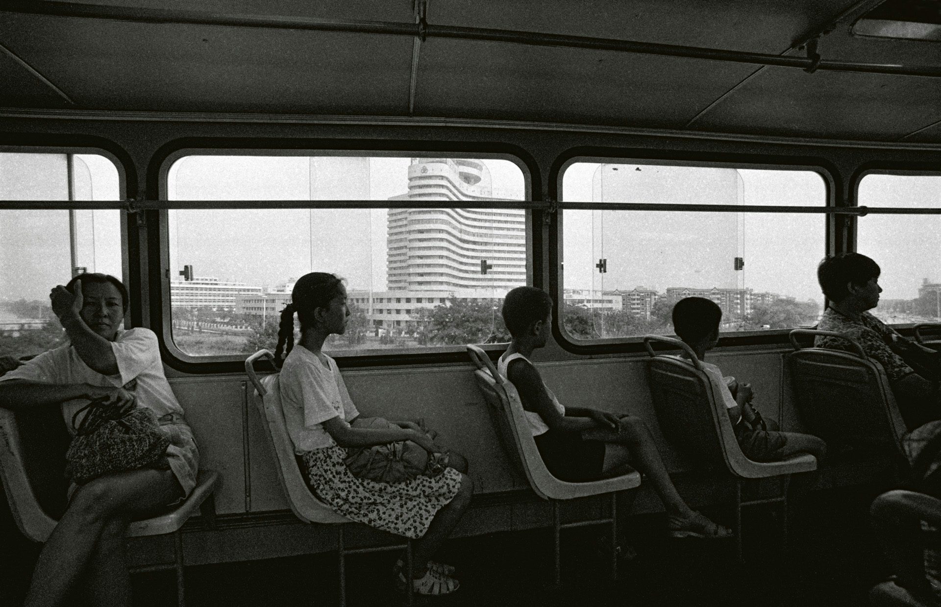Black and white photo by Mo Yi showing passengers sitting in a single row of bus seats, taken from the other side of the bus