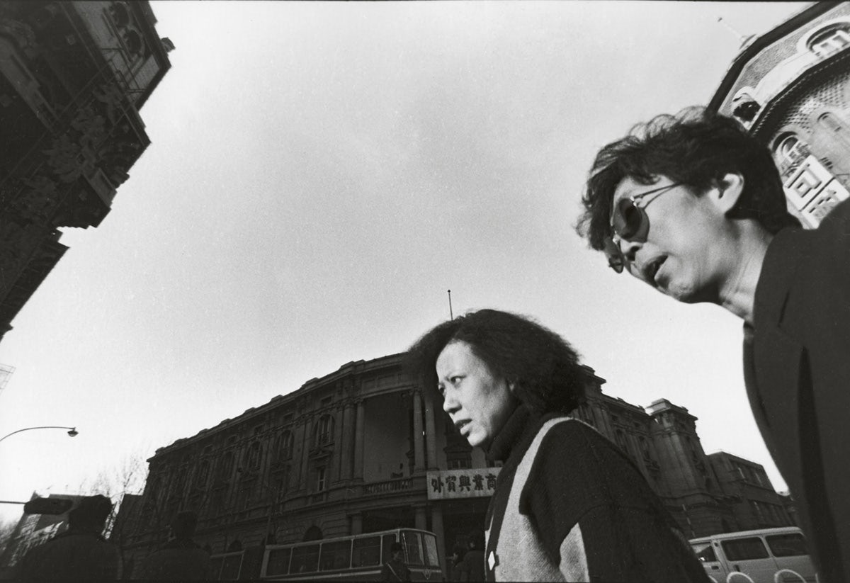 Black and white photo by Mo Yi taken from a low perspective showing passersby, one of which is wearing sunglasses, against a backdrop of buildings and sky