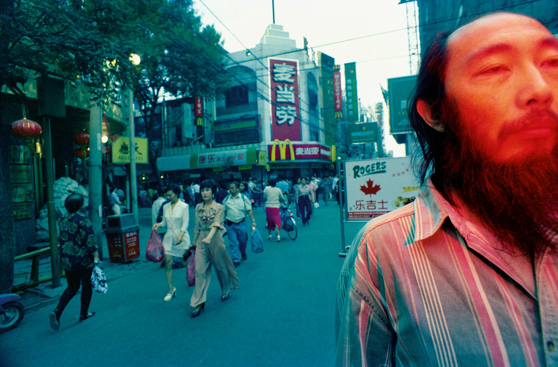 Photo by Mo Yi featuring the artist's face in the foreground and a busy street of stalls and passersby in the background