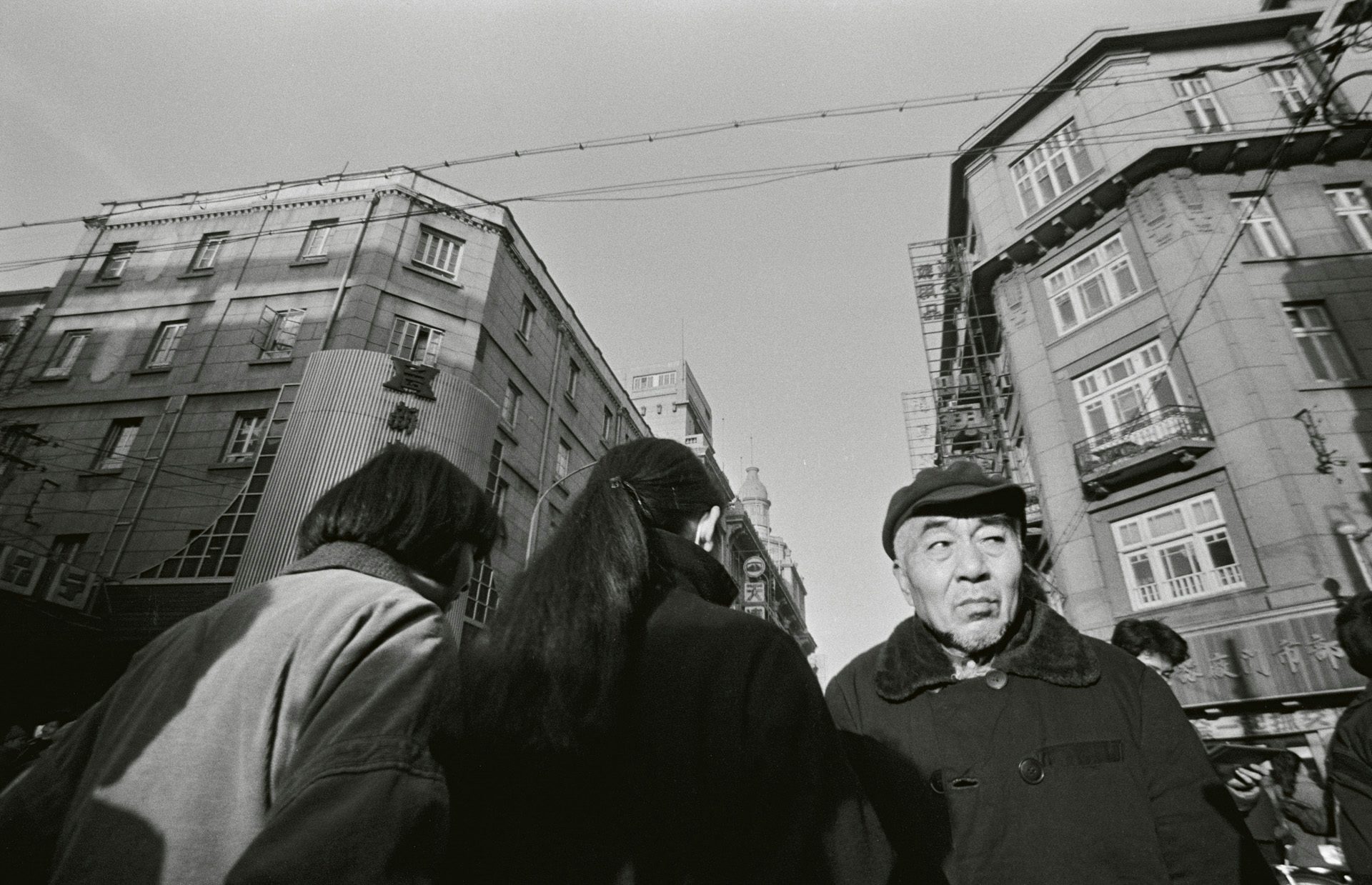Black and white photo by Mo Yi showing people walking in a city street taken from a lower perspective, showing buildings in the background