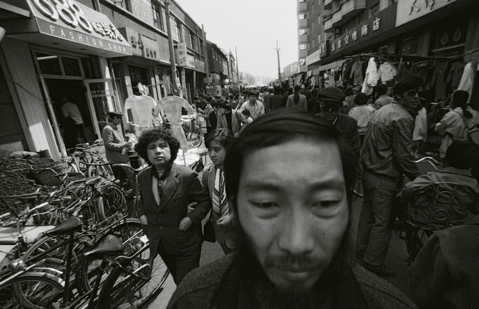 Black and white photo by Mo Yi featuring the artist's face in the foreground and a busy street of stalls and passersby in the background