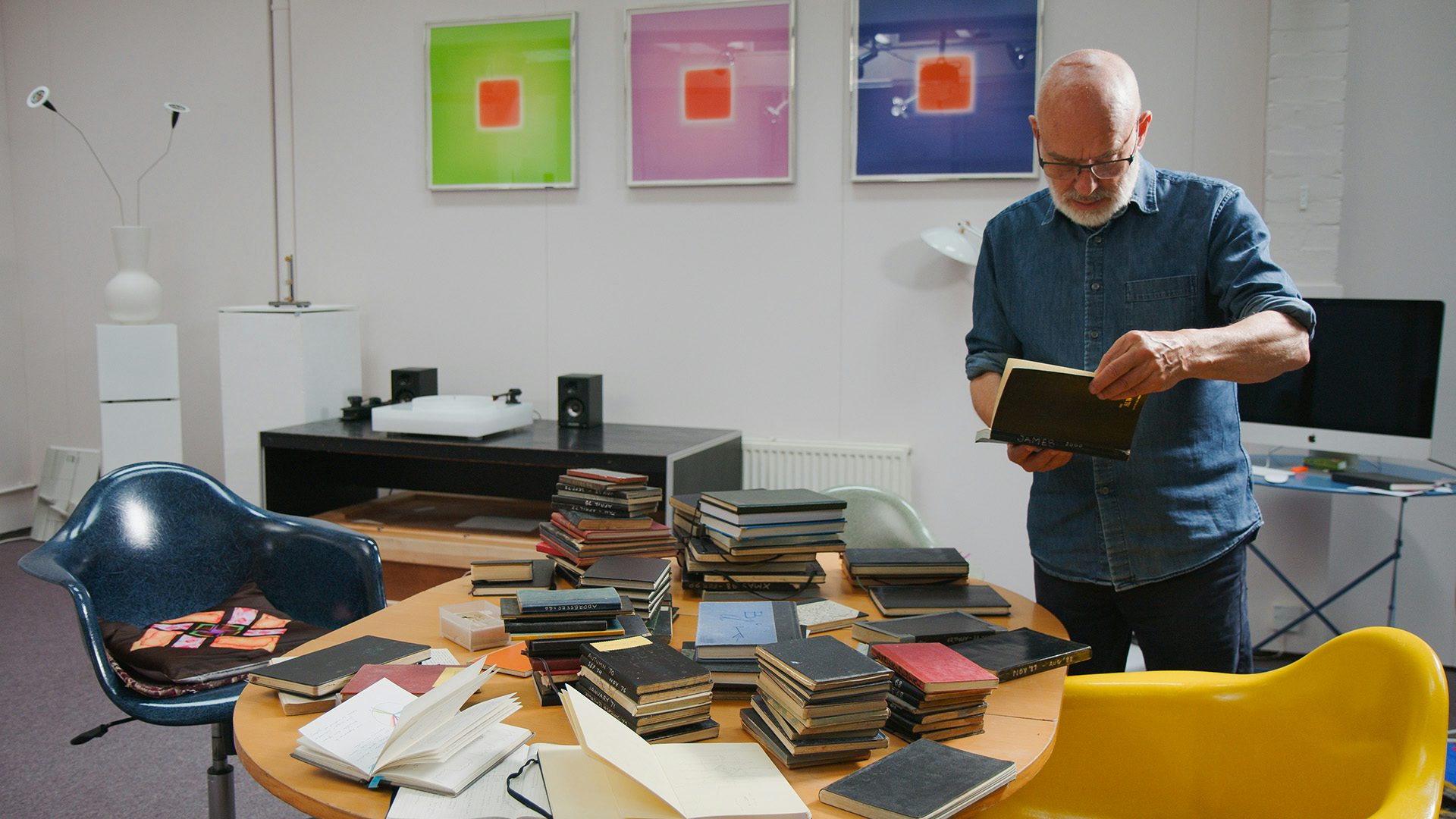 Briano Eno reading a notebook standing over a table filled with stacks of notebooks, and three square abstract artworks in the background