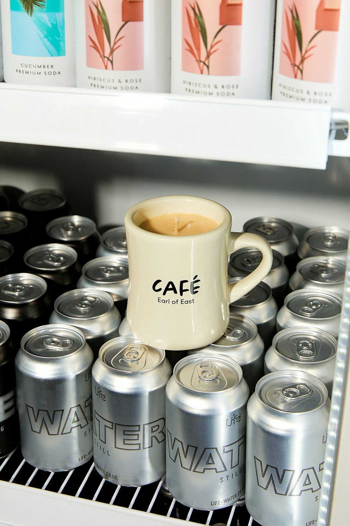 A beige mug labelled 'cafe' on top of a shelf full of silver drinks cans