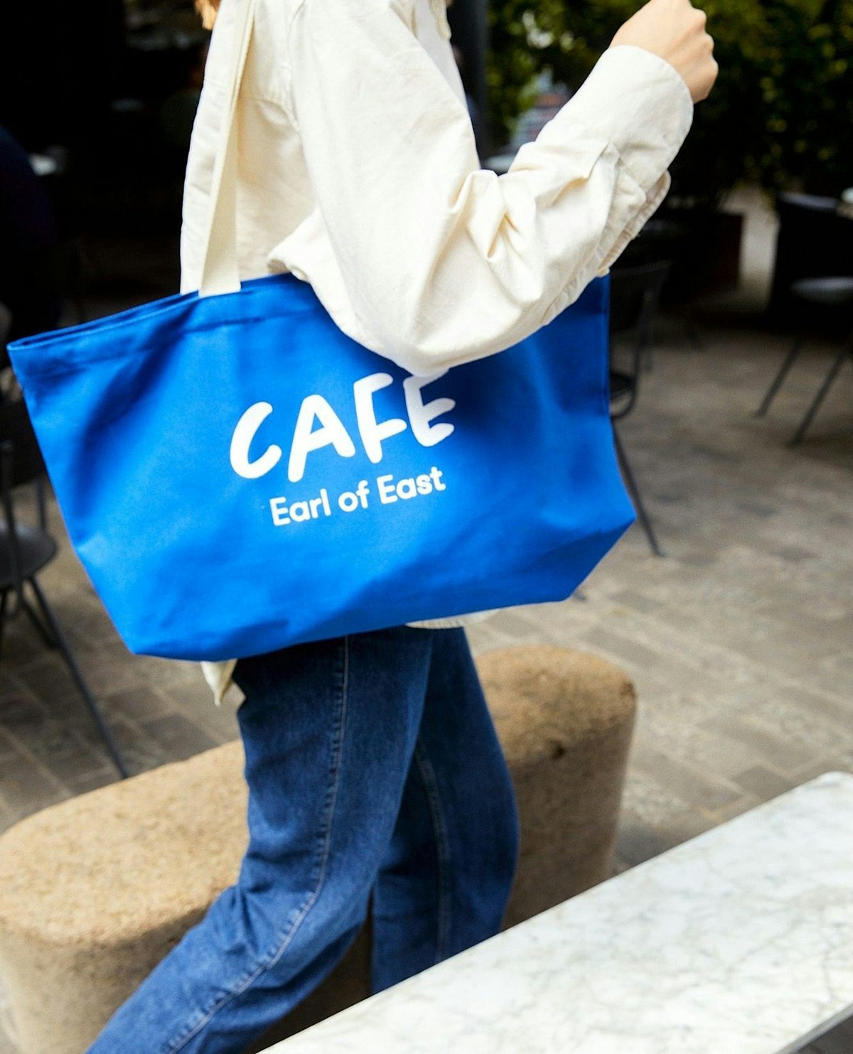 A person wearing jeans, a light shirt and a bright blue tote bag labelled 'cafe'