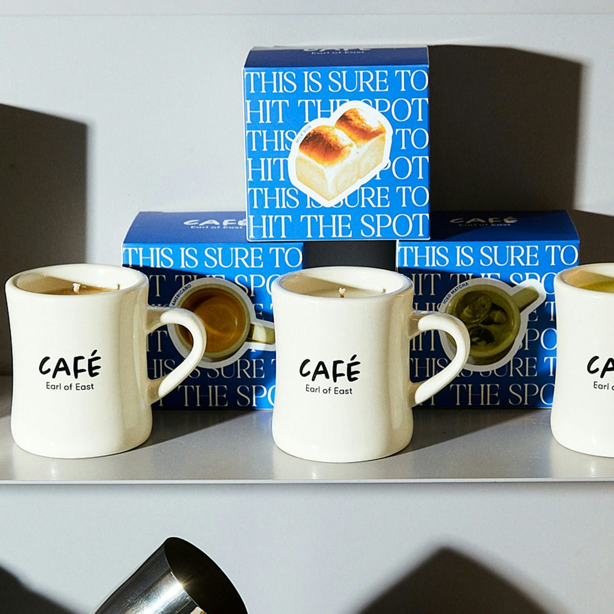 Three beige mugs labelled 'cafe' on a shelf surrounded by packaging and glassware