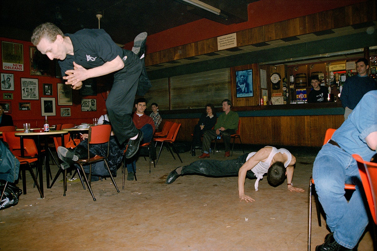 Two people dancing in a bar, with several people sat around watching. One person is jumping in the air, the other person dancing on the floor