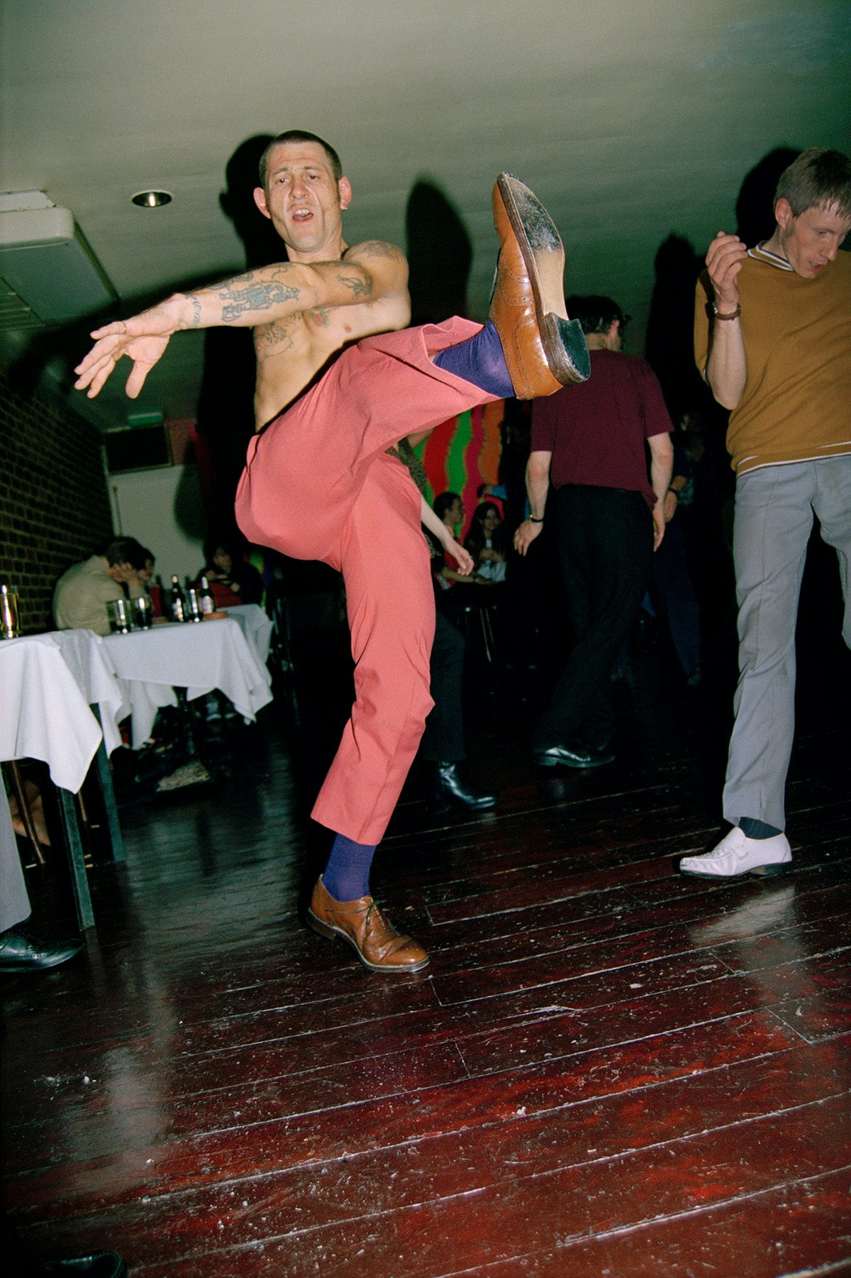 A topless person wearing salmon coloured trousers dancing with a leg kicked up in the air