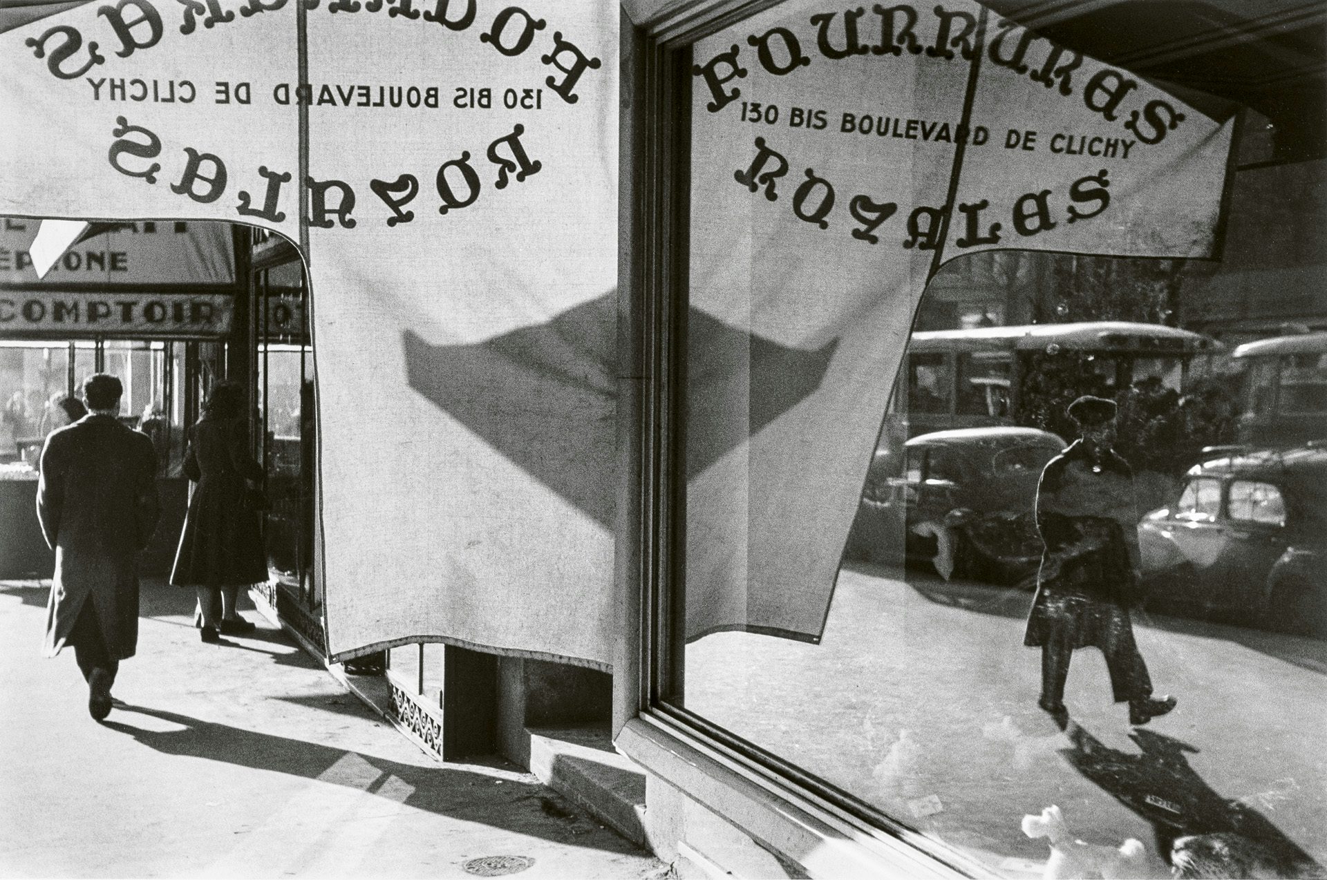 Black and white photo by Louis Stettner of a person walking in the street and another person walking in the opposite direction shown in the reflection of the same scene