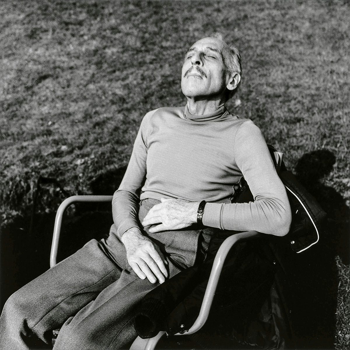 Black and white photo by Louis Stettner of a person with closed eyes and a thin moustache reclining in a chair in a park