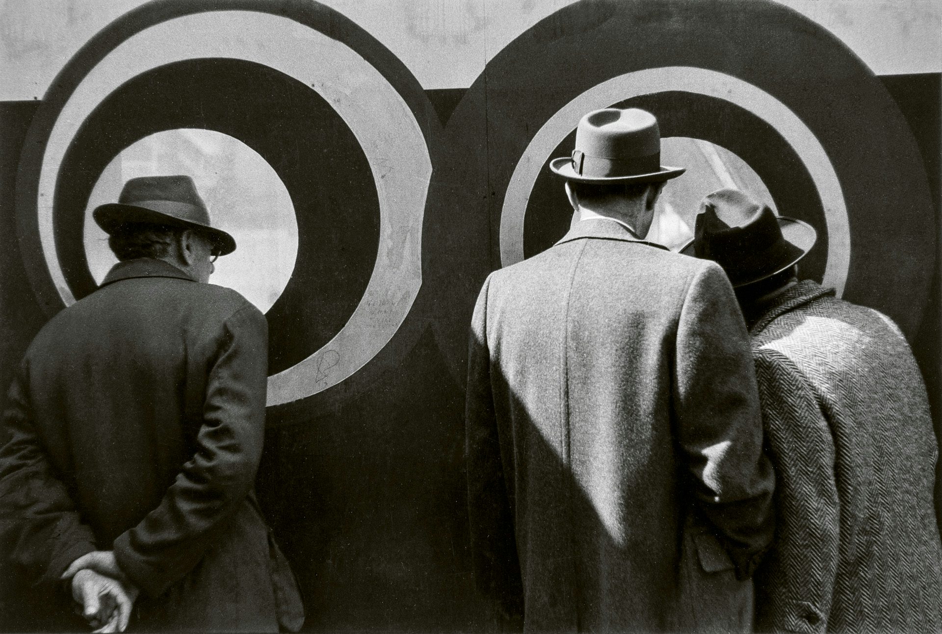 Black and white photo by Louis Stettner of people in overcoats and hats with their backs turned to the camera looking at concentric circles on a wall behind them