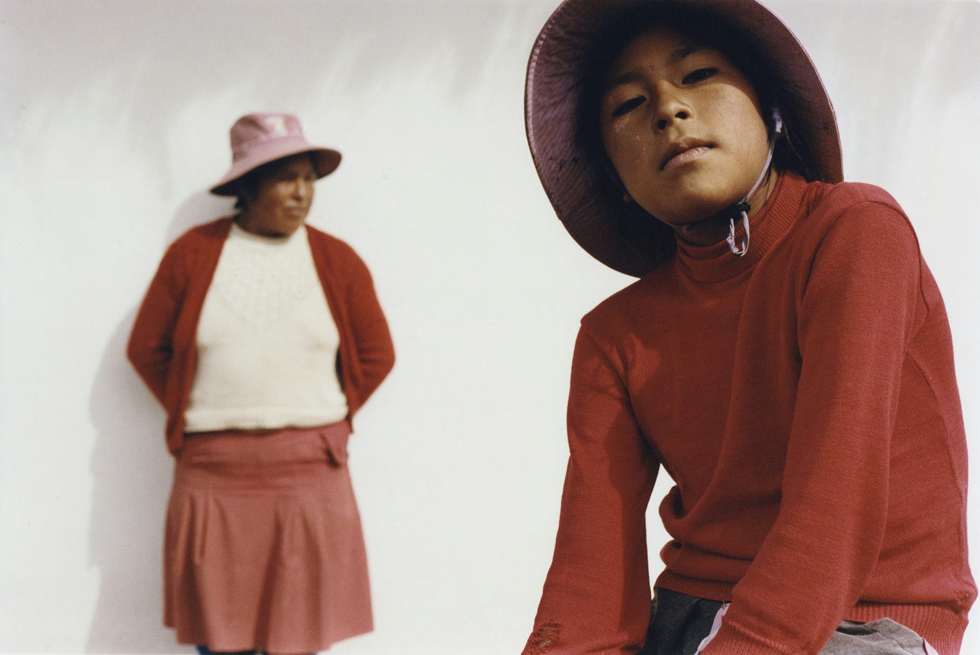 Photo by Ana Margarita Flores of two people against a white backdrop, wearing white and deep red outfits and pale purple headdress. One person in the rear is out of focus, the other person in the foreground looks towards the camera
