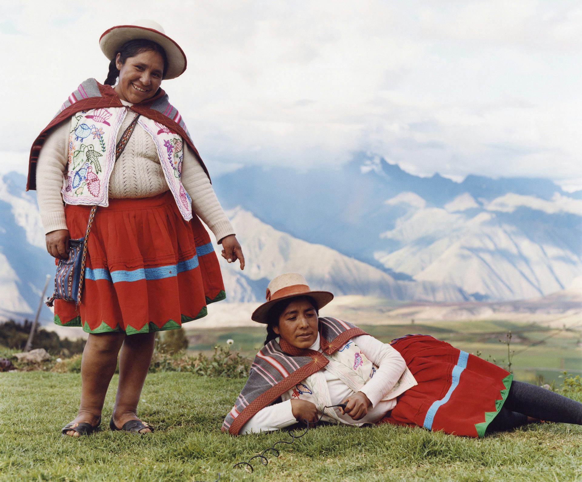 Photo by Ana Margarita Flores of two Peruvian women wearing embroidered waistcoats and red skirts. One person is standing and smiling, the other person is lying on the grass and holding a shutter release. In the background are misty mountains