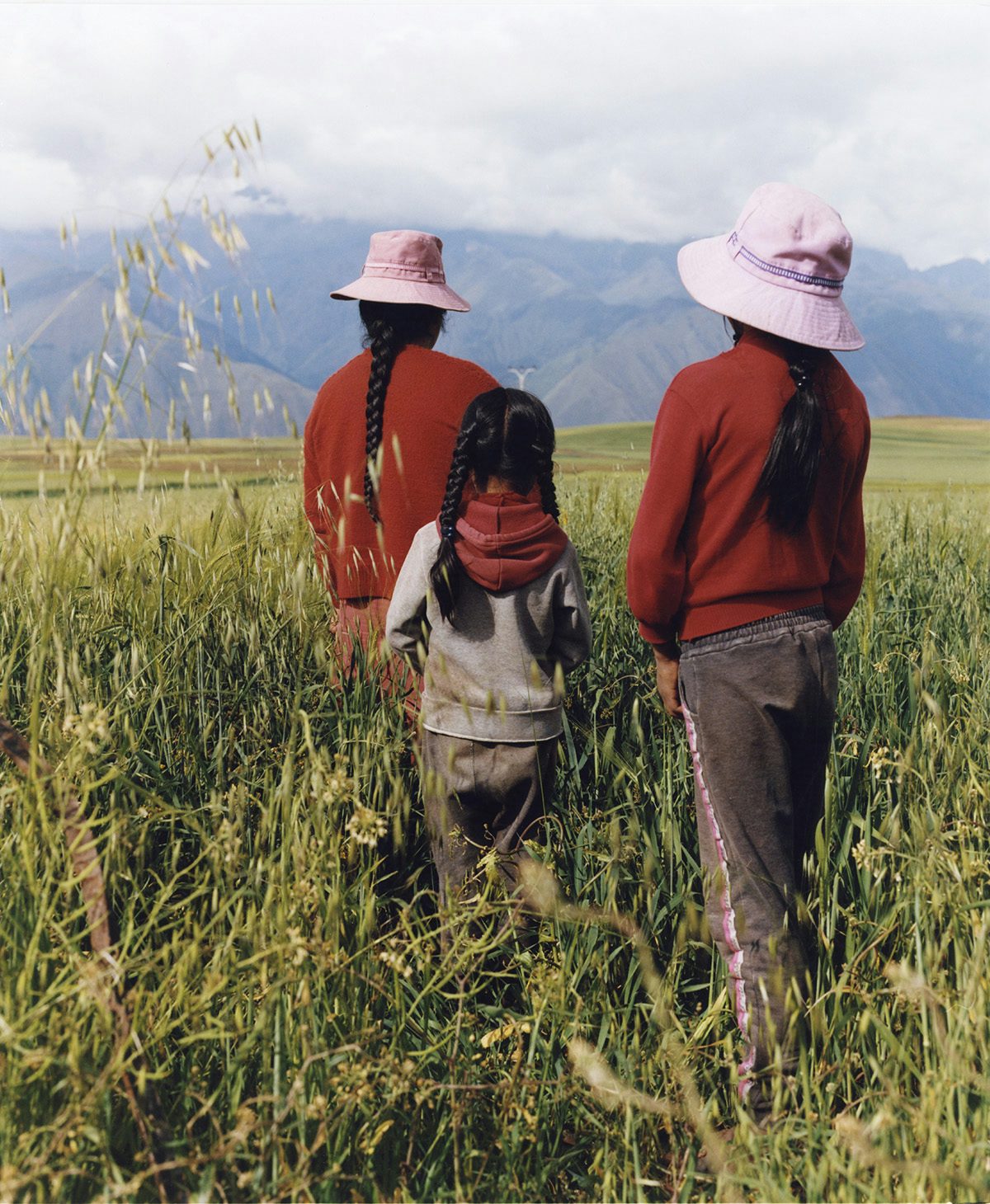 Photo by Ana Margarita Flores of three people, two of them wearing crimson long-sleeved shirts and pale purple headwear, with their backs facing the camera as they walk through a field towards misty mountains in the background