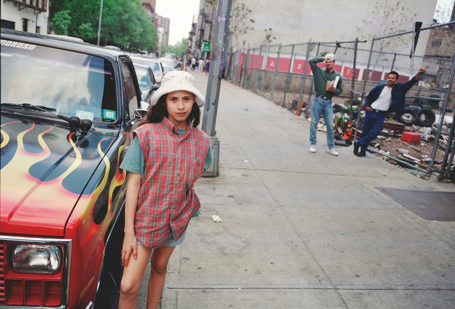 A girl wearing a white hat and a checkered sleeveless shirt and a t-shirt underneath, standing in front of a city street with two people hanging out in the background and a a black car with flames painted on the bonnet