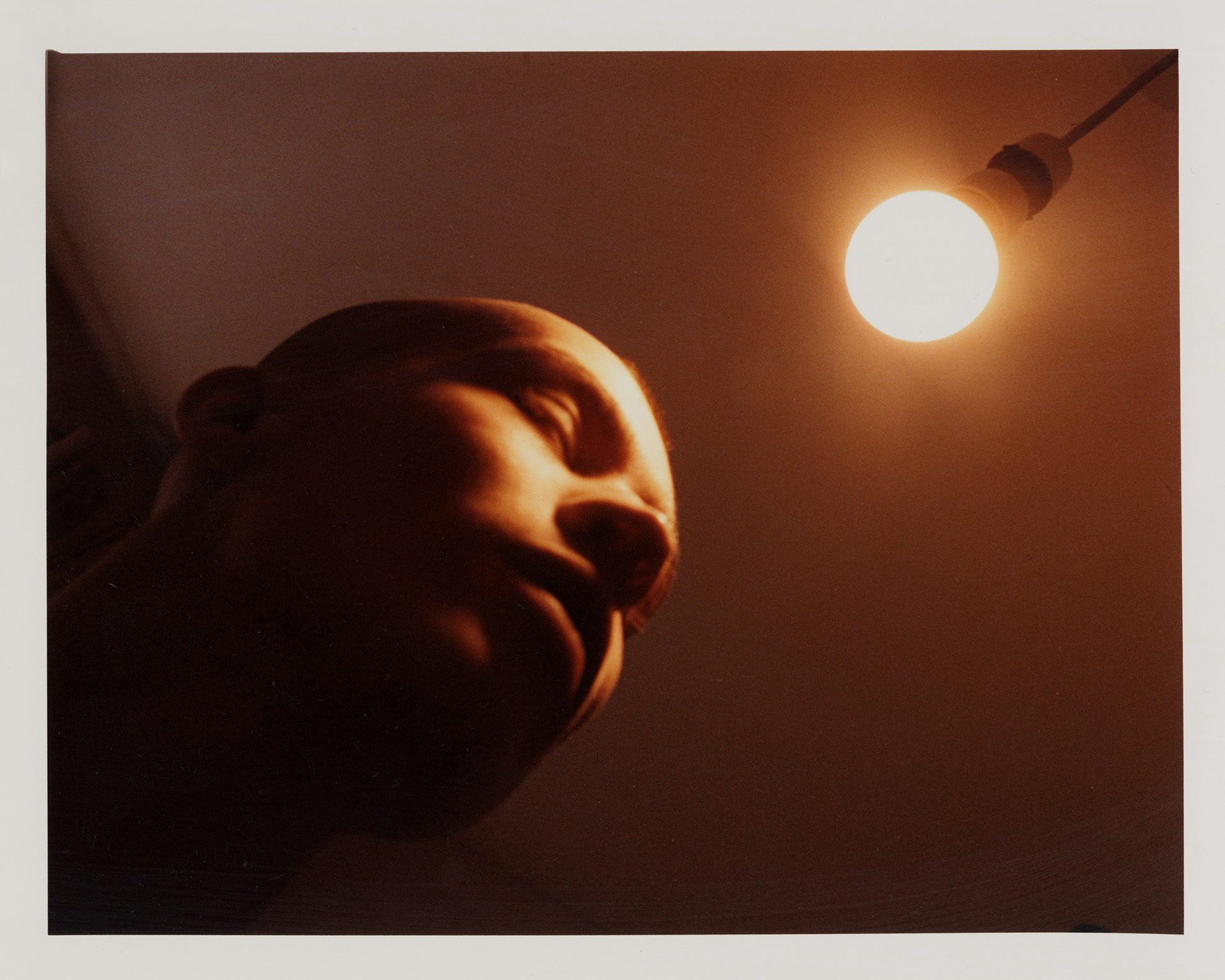 Orange hued photo of a person with their eyes closed leaning towards a glowing ceiling light bulb