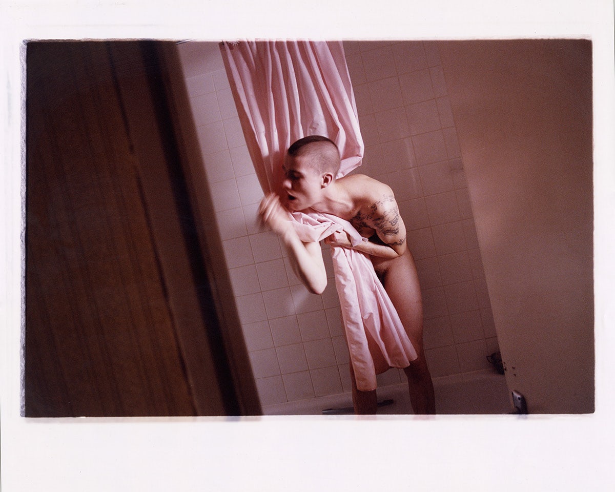 A person wearing no clothes standing in a bathtub clutching a pink shower curtain