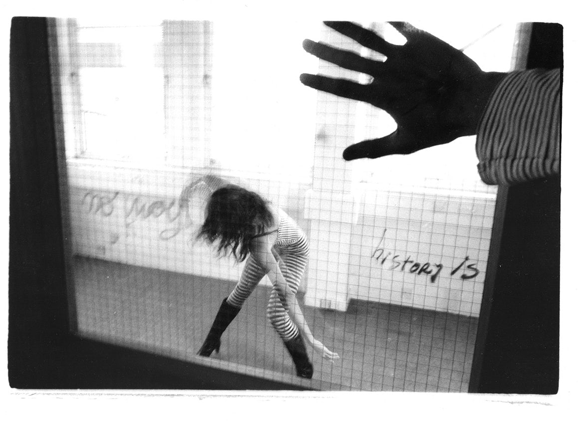 Black and white photo of a hand held up to a window through which a person contorting their body in a corridor can be seen