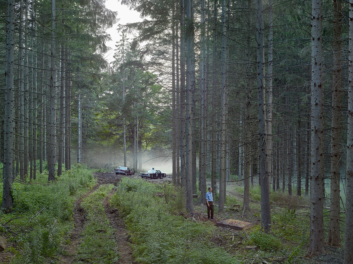Eerie woodland scene with two seemingly abandoned cars in the background and a person in the foreground staring in the distance