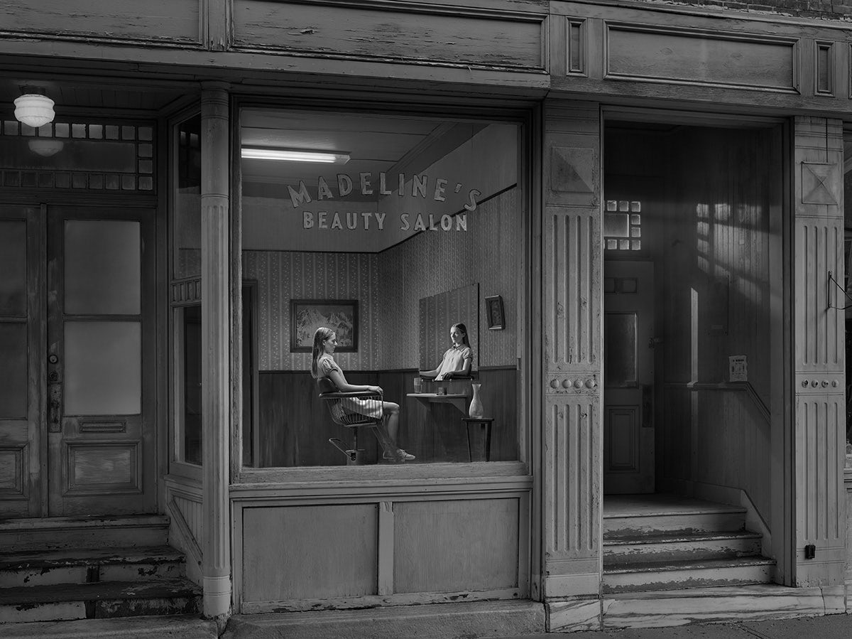 Black and white photo by Gregory Crewdson of a person sat in a beauty studio opposite their reflection