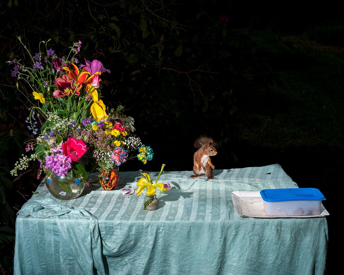 A tabletop covered in a pale blue tablecloth, on top is a vase of bright flowers next to a squirrel