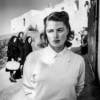 Black and white photo of Ingrid Bergman wearing a sweater and collared shirt, with three people in the background wearing dark clothes and headscarves looking at her