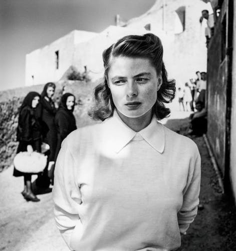 Black and white photo of Ingrid Bergman wearing a sweater and collared shirt, with three people in the background wearing dark clothes and headscarves looking at her