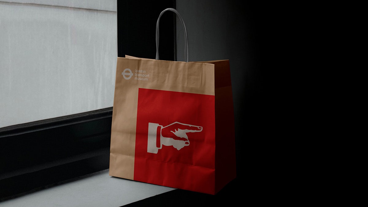 A London Transport Museum branded brown paper bag featuring a red square and a pointing finger icon