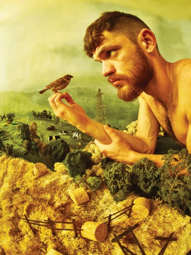 Photo by Tim Walker and Shona Heath of a person with short hair and a beard and bare chested, with a sparrow resting on their hands, with a crafted miniature farm in the background