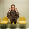 Zalando campaign photo of Willem Defoe crouching on a row of yellow chairs in a pale green hued room