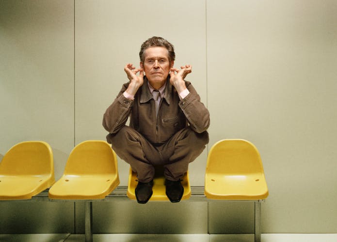 Zalando campaign photo of Willem Defoe crouching on a row of yellow chairs in a pale green hued room