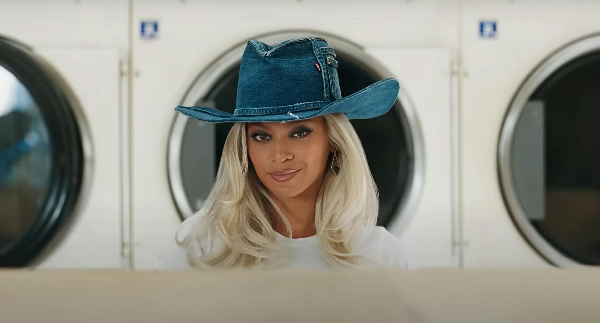 Still from a Levi's advert showing Beyoncé with blonde hair and wearing a raw denim cowboy hat standing in front of a washing machine in a laundrette