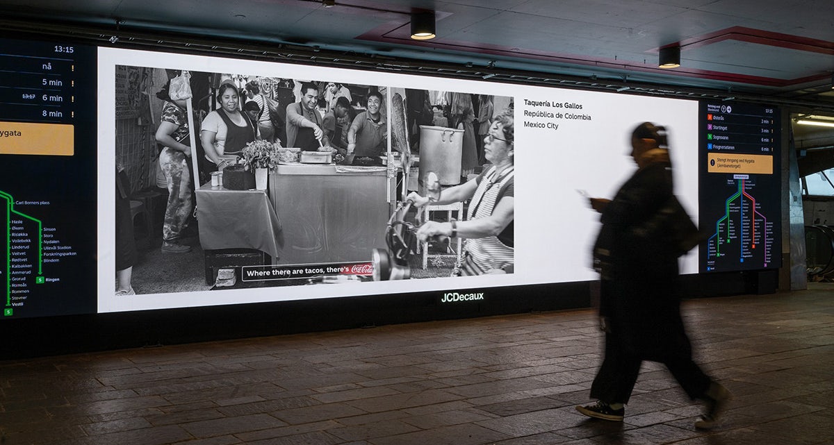An outdoor ad featuring a black and white photo of a taco stall with a crate of Coca-Cola next to the stall highlighted in red