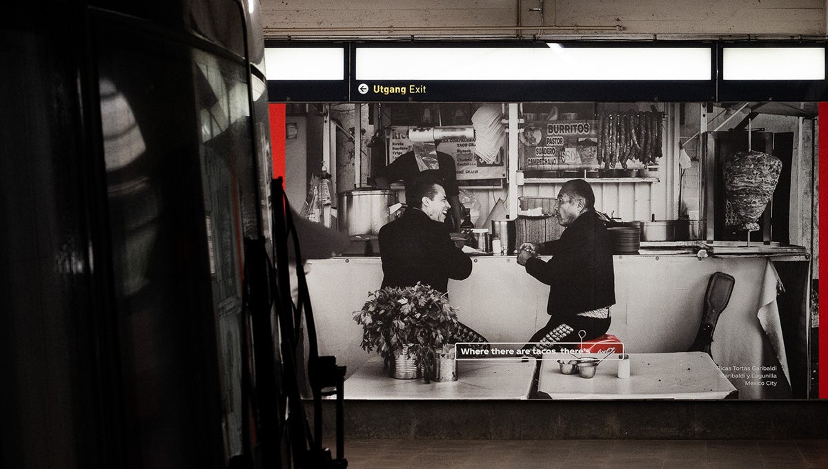 An outdoor ad featuring a black and white photo of a taco stall with a crate of Coca-Cola next to the stall highlighted in red