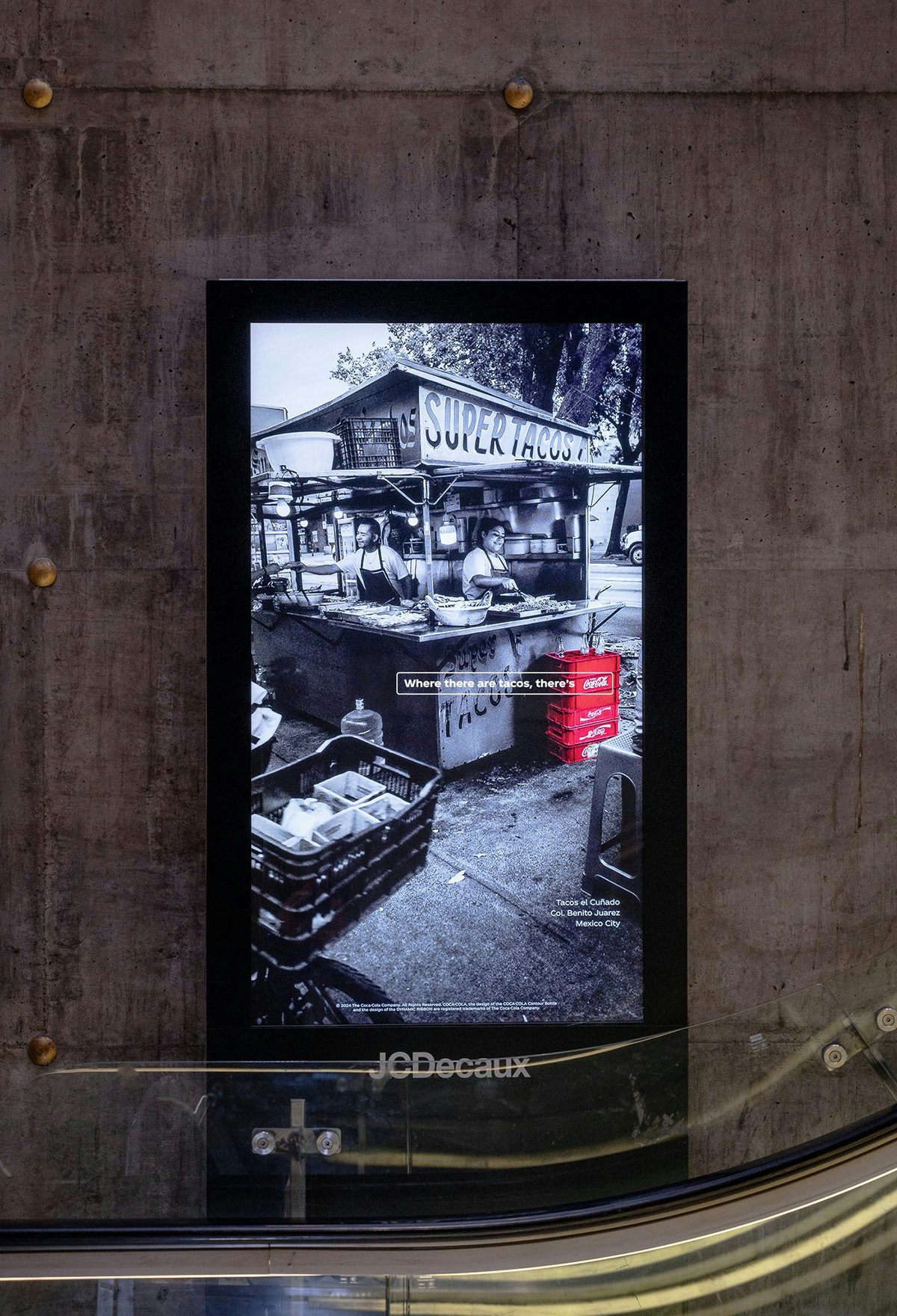 An outdoor ad featuring a black and white photo of a taco stall with a crate of Coca-Cola next to the stall highlighted in red
