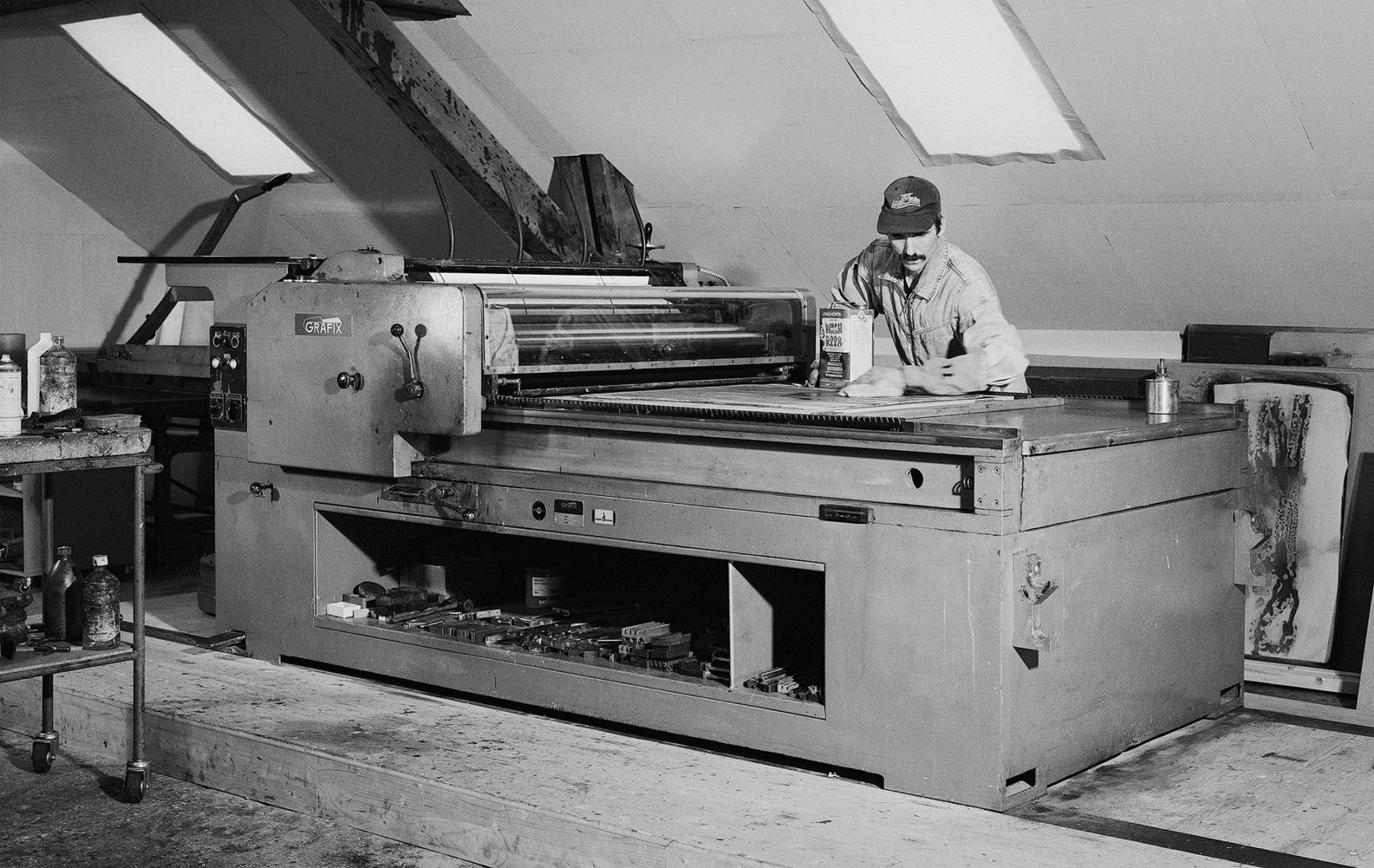 Black and white photo of poster designer Dafi Kuhne in a studio in front of a large press