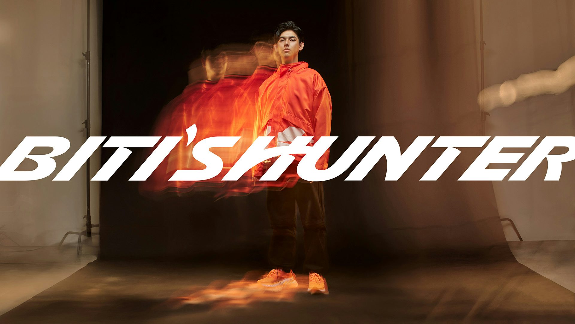 Biti's Hunter wordmark in white lettering with anglar details, laid over a motion blurred photo of a person wearing a bright orange windbreaker jacket and trainers