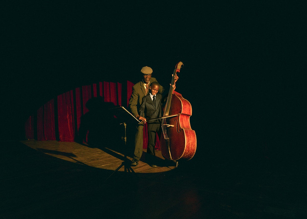 Photo by Carlos Idun-Tawiah showing an adult showing a child how to play the double bass on a low-lit stage with red curtains in the background