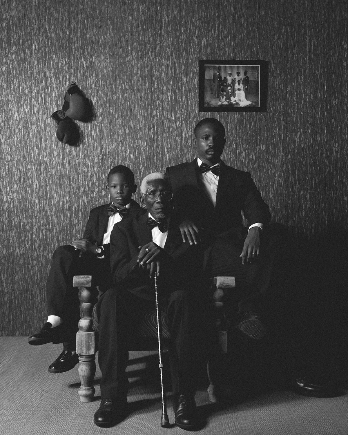Black and white photo by Carlos Idun-Tawiah showing three generations of men sat on and around an armchair looking into the camera
