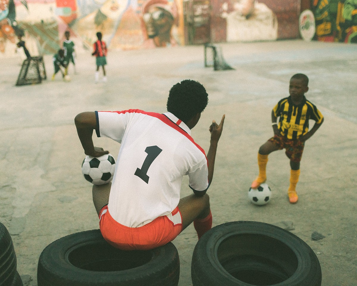 Photo by Carlos Idun-Tawiah showing a person wearing a football top with their back to the camera and sat on top of tyres holding a football under one arm and pointing at a child standing in a playground in front of them