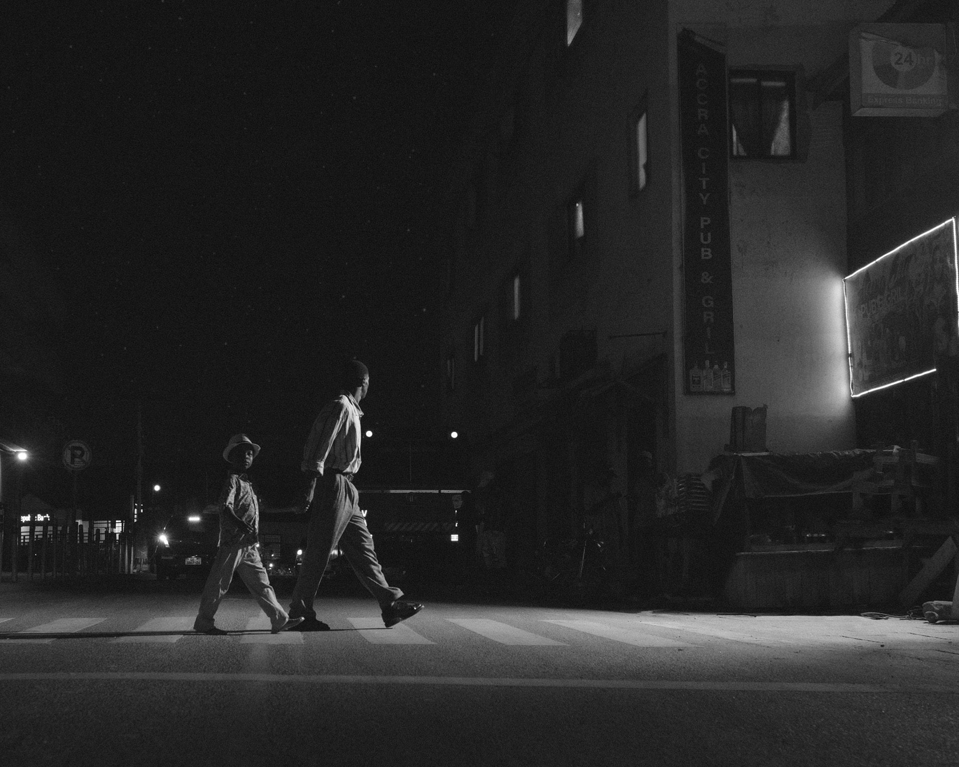 Black and white photo by Carlos Idun-Tawiah showing a man and a child crossing the road in a low-lit street