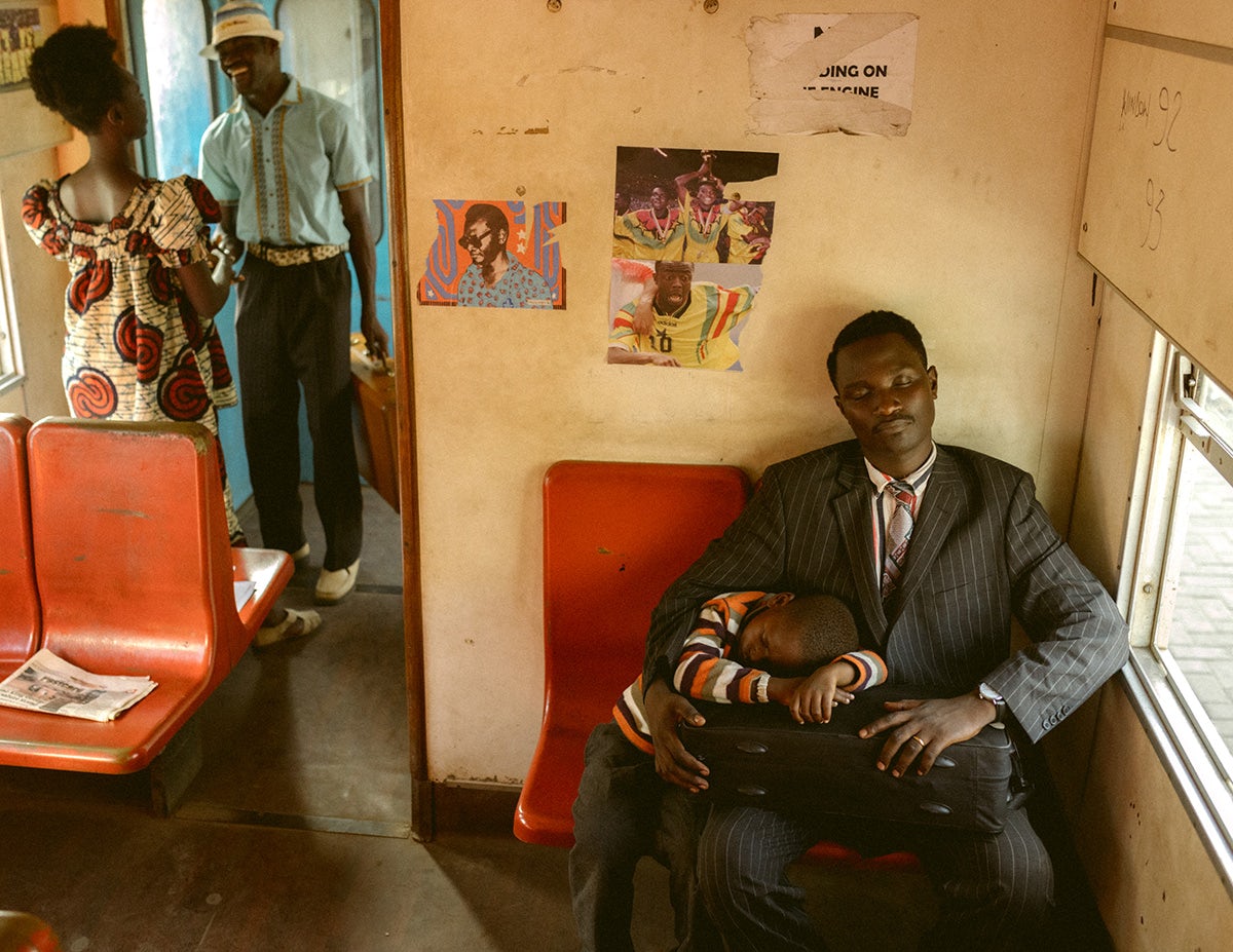 Photo by Carlos Idun-Tawiah showing an adult on a train wearing a suit with a child resting underneath their arm