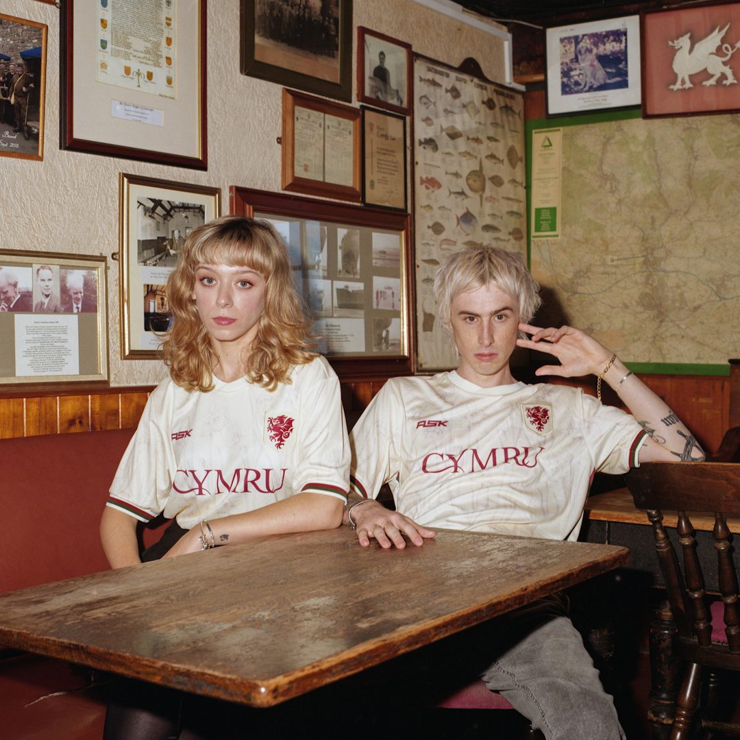 Photo by Callum O'Keefe showing two young people sat in an old fashioned pub wearing white football shirts labelled 'Cymru'
