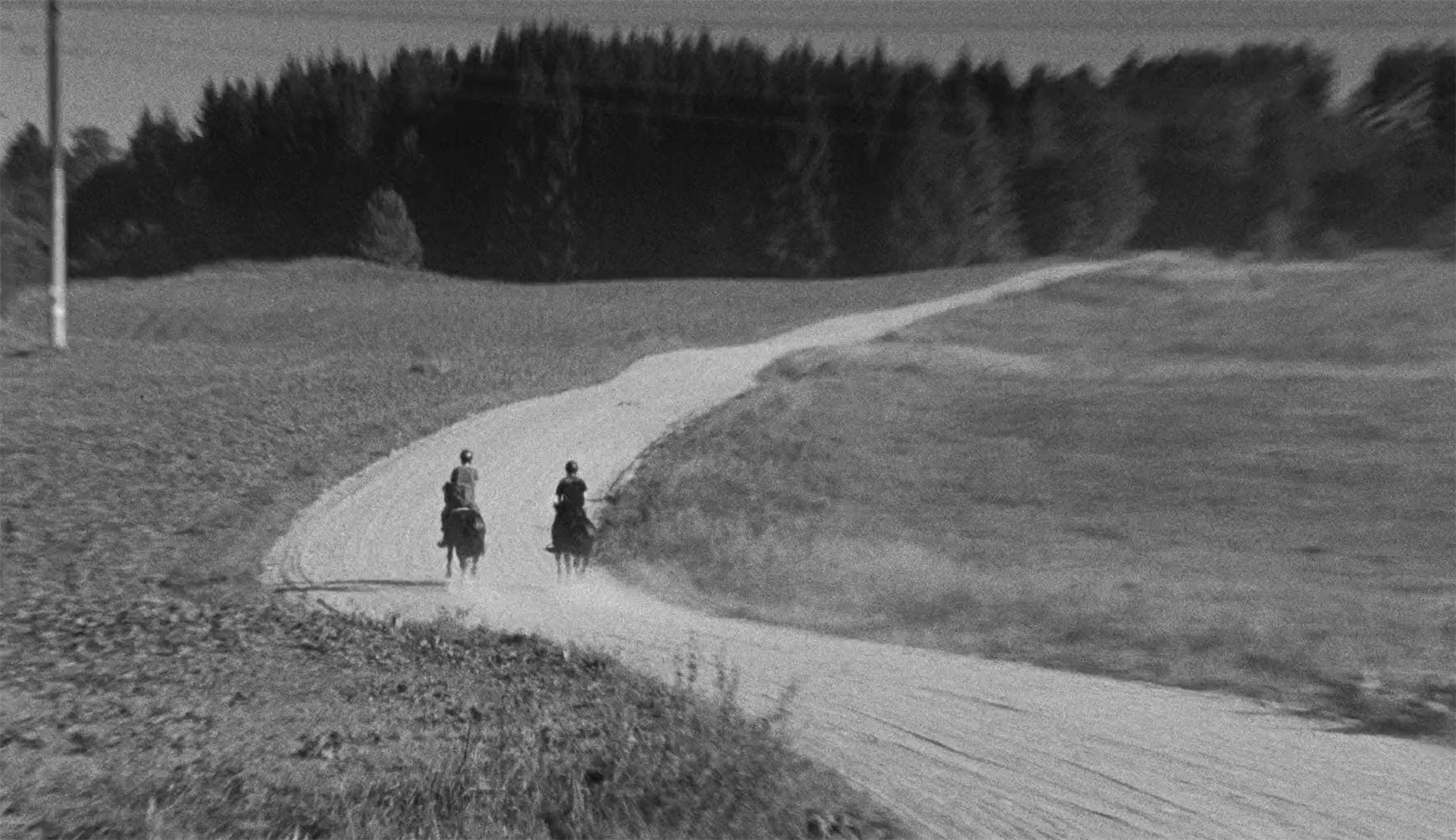 Black and white still showing two people riding horses on a dusty track leading towards a forest