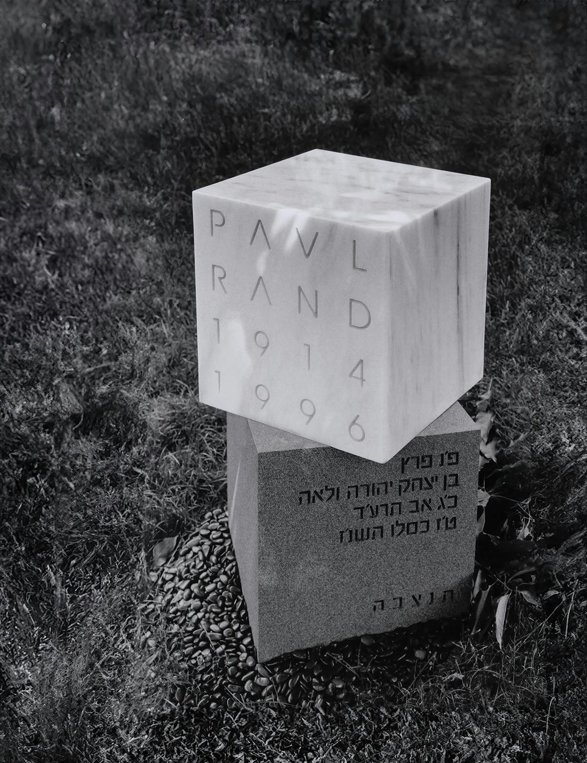 Black and white photo of Paul Rand's headstone designed by Fred Troller, involving a stack of two cube structures with his name and the dates 1914 and 1996 on the top cube, and Hebrew inscription on the bottom cube