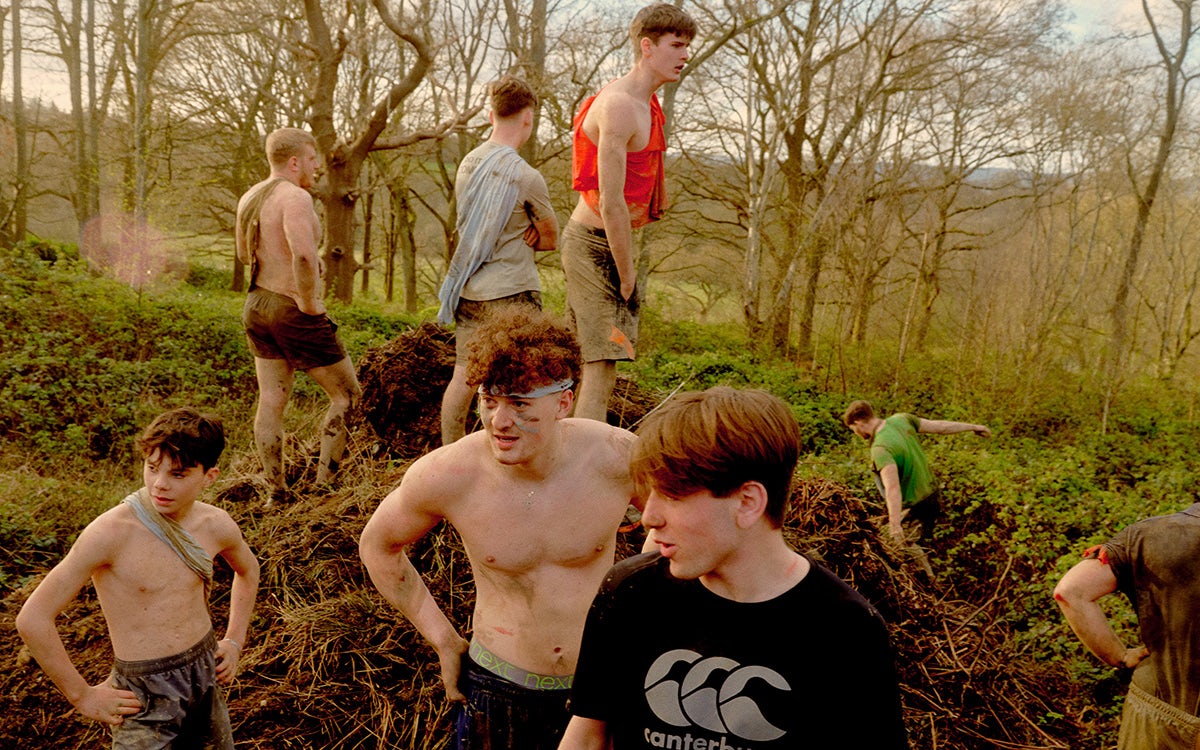 Young men in shorts covered in mud while running through a field next to a woodland