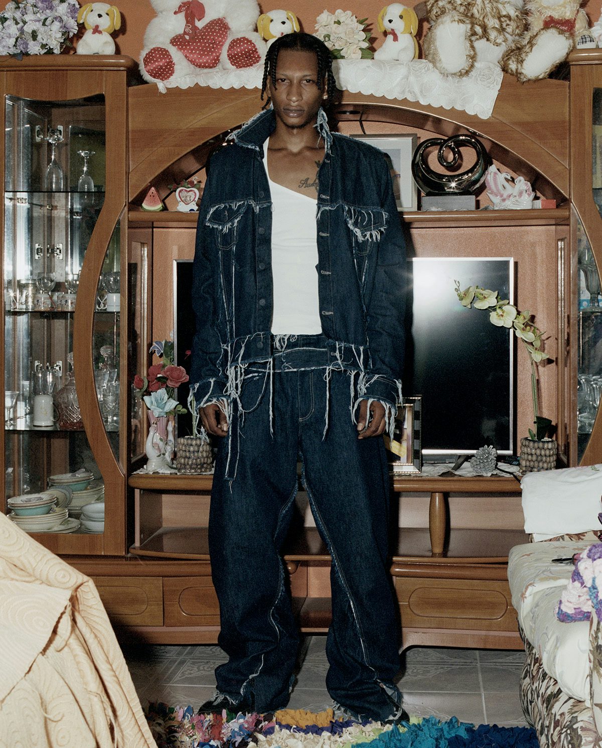 Photo by Kwabena Sekyi Appiah-nti of a man wearing a matching frayed denim jacket and trousers standing in a living room with a TV and large wooden dresser in the background