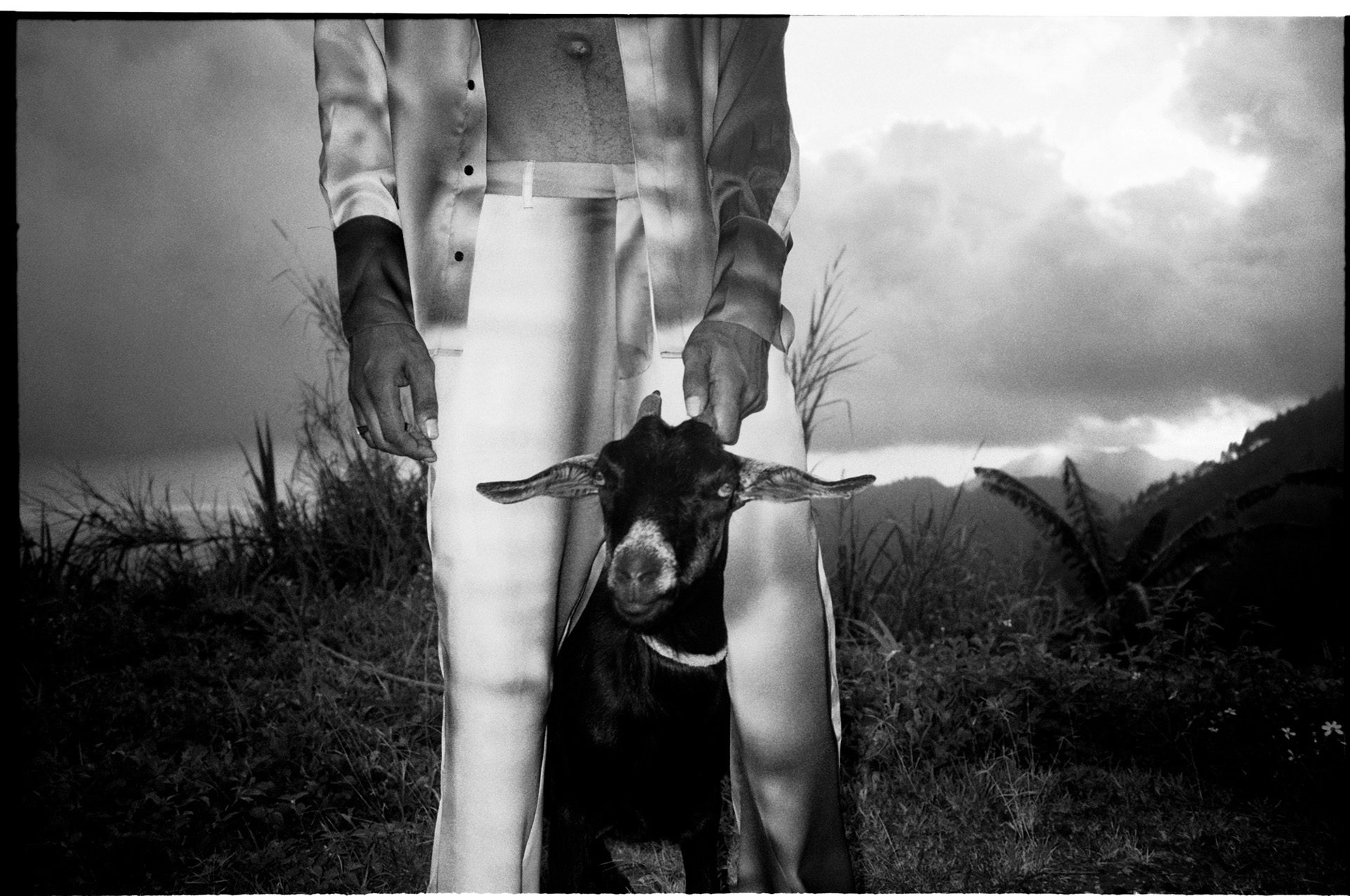 Black and white photo by Kwabena Sekyi Appiah-nti of a person wearing silky textured trousers with a goat held between their legs, standing against a dramatic landscape