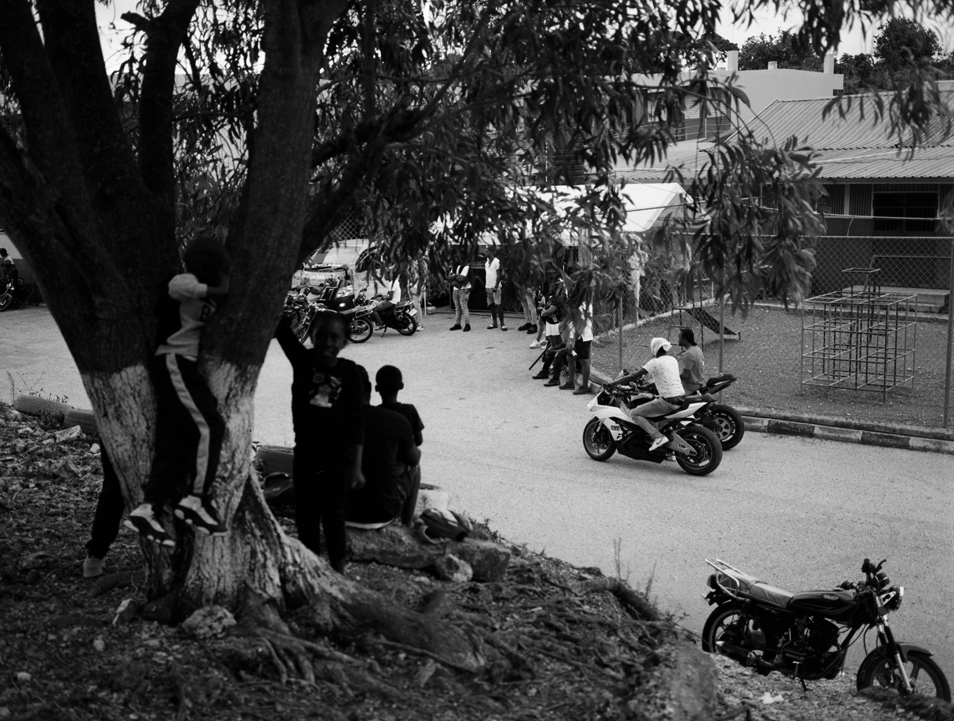 Black and white photo by Kwabena Sekyi Appiah-nti taken from afar showing people hanging out around a street where two people are riding motorbikes alongside a large tree