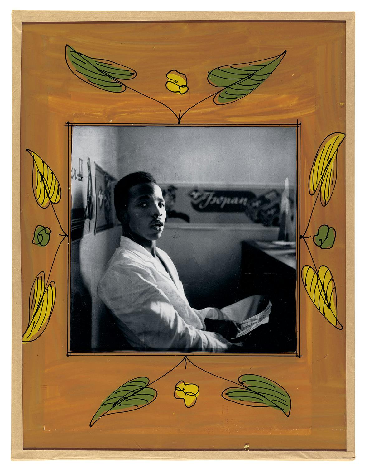 Black and white photo of a person sitting at a table, which is bordered by a mustard coloured painted frame with green and yellow leaves