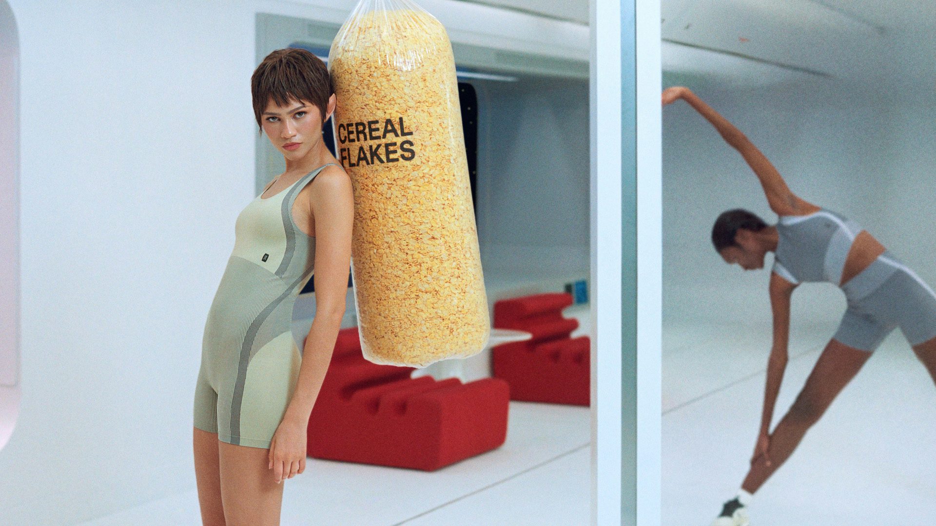 Still from On's Zone Dreamers ad featuring Zendaya wearing sportswear and leaning on a clear punching bag filled with cereal and labelled cornflakes, with another person stretching behind her